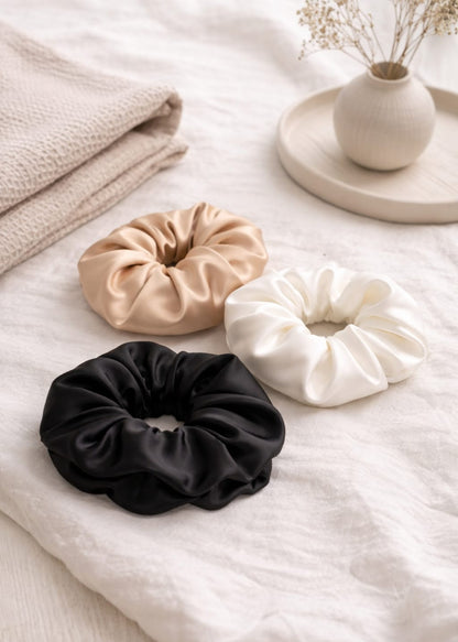 Three Silk scrunchies (beige, black, and white) on a soft fabric surface.
